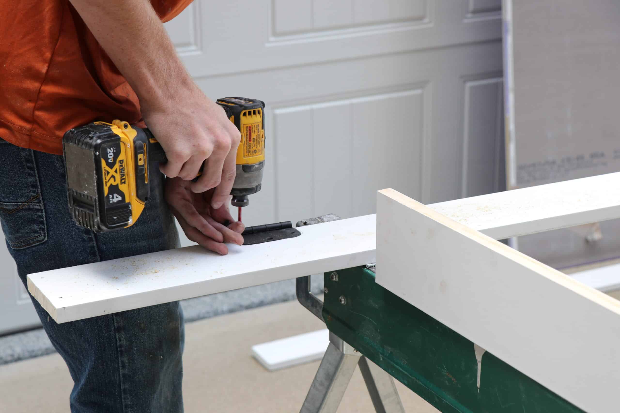 Worker in an orange shirt holds a yellow DeWalt cordless drill, drilling into a white wooden board on a worktable at a construction site